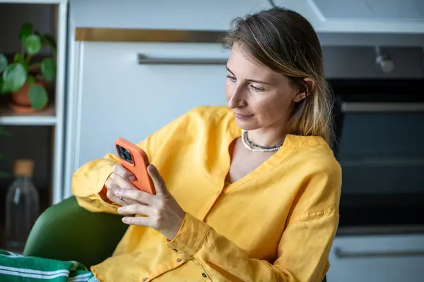 Relaxed Swedish woman reading online message, scrolling social media news at home. Scandinavian long-haired female using smartphone apps sitting at chair at kitchen. Digital addiction concept.