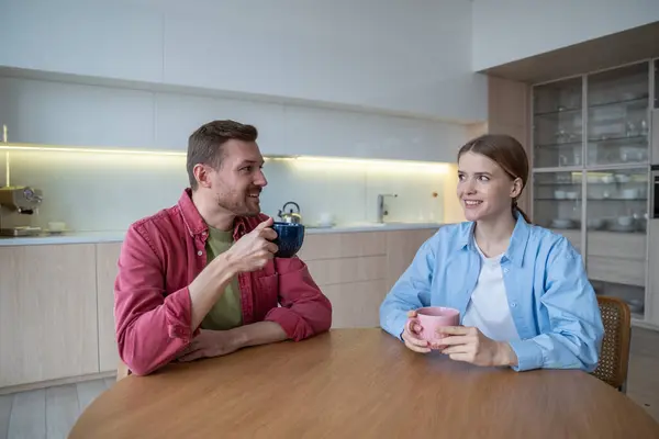 Pleased couple love at kitchen table, holding mugs coffee in hands have pleasant conversation. Young family man and woman smiling, looking each other with warmth and understanding talk over cup tea.