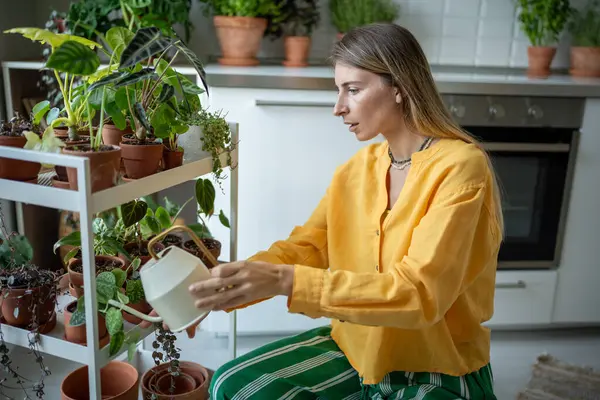 stock image Serious young florist woman in yellow linen shirt waters sprouts indoor plants in flower pots with metal watering can. Home gardening, love of houseplants and care, mental green hobby concept