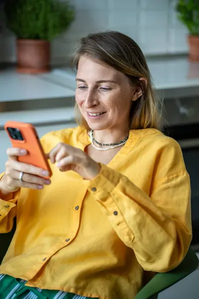 stock image Happy woman housewife with phone chatting with friends in online messenger sitting in chair in kitchen. Joyful Swedish female in casual yellow shirt flipping through social media feed in smartphone 