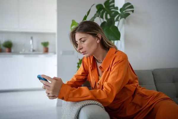 Lonely fatigued woman sitting on sofa holding smartphone sadly looking at screen scrolling web social media. Tired middle aged scandinavian female waiting message or call feeling upset, frustration.