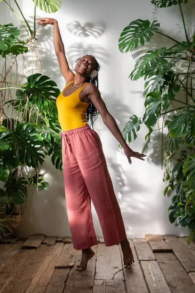 アフリカ系アメリカ人の若い女性は 緑豊かな家の植物のなかで歌にリラックスして 楽しい瞬間 ヘッドフォンで裸足で踊るケアフリーの黒人女性 木製の床で音楽に没頭した純粋な喜び — ストック写真
