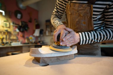 Closeup of sanding wood with orbital sander at workshop. Man carpenter polishes wooden seat of a future chair with electric sander. Carpentry workshop. Furniture production clipart