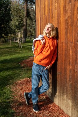 Happy guy basks in warm rays of sun while walking outdoor. Cheerful long-haired blond man leaning effortlessly against the wooden wall with eyes closed in bliss savouring every moment of serenity joy. clipart
