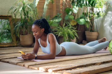 Contented african american woman reclining on fitness mat with smartphone in hand, pausing after home exercise session surround by houseplants. Sported black girl lying on wooden floor with cellphone. clipart