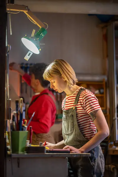 stock image Focused woman carpenter writes down list of materials need to produce handmade wood products in carpentry workshop. Small business. Interested in sales craftworker makes an estimate on wood production