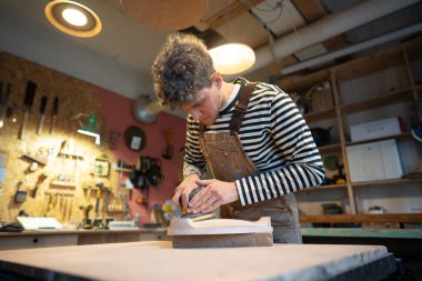 Sanding wood with orbital sander at workshop. Concentrated man carpenter polishes wooden seat of a future chair with electric sander. Carpentry workshop. Furniture production clipart