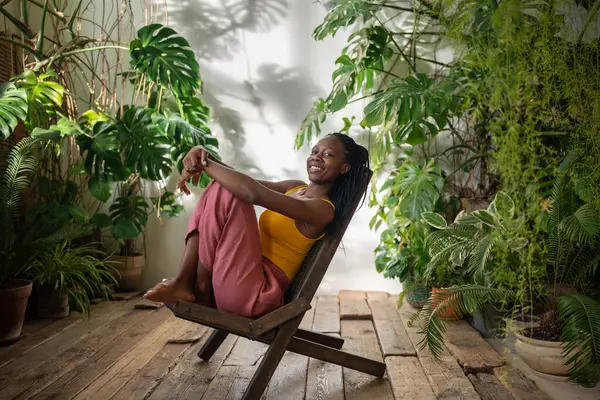 stock image Joyful happy african american woman rest on chair surround by tropical houseplants enjoy calmness in sunny day look at camera. Playful pleased black young female smiling sit in room with indoor plants