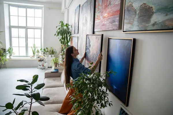 Woman painter hanging newly painted modern framed painting on wall in bright room art workshop with indoor plants. Actual solutions by decoration in contemporary interior design. Artists cosy home