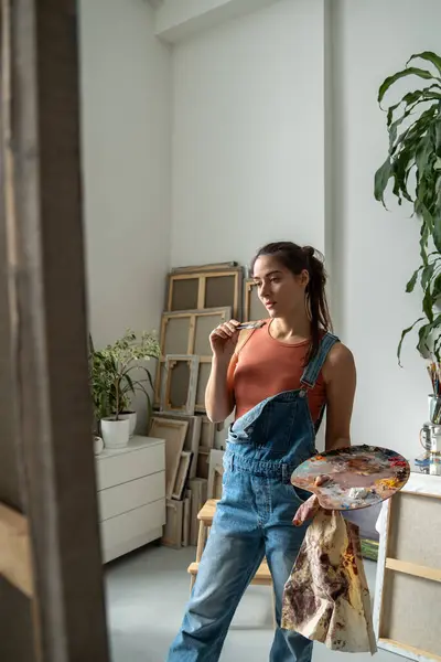 Stock image Thoughtful woman artist looks on canvas on easel pondering about creative idea for picture holds oil colour palette, paintbrush standing in art studio. Small craft business, stress relieve interests.