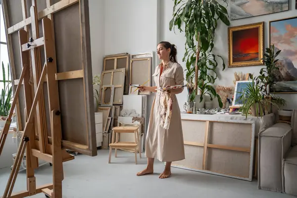 stock image Thoughtful artist woman with inspiration paints picture, pondering ideas, holding in hands palette with oil paints in front of easel with canvas. Creative female standing barefoot in cozy art studio. 