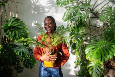Mutlu Afrikalı Amerikalı kadın bitki sever, sarı tencerede sicaları tutuyor güneşli bir günde kameraya gülümsüyor. Çiçekçi dükkanının neşe dolu, başarılı zenci kadın sahibi portresi