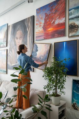 Kadın sanatçı duvara yeni bir resim astı. Kız evdeki kompozisyonu değerlendiriyor. Ev iç tasarımına ciddi yaklaşım, apartmanı dekorasyonlarla doldurmak yaratıcı bir ortam yaratmak.