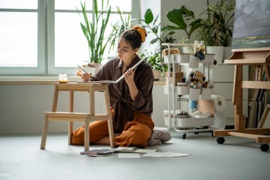 İlgili kadın ressam çizim kitabı fırçasıyla resim çiziyor. Sanat stüdyosunda yerde bitkilerle oturuyor. Resim sehpası yakınlarındaki tasarım düzeni için ev sanatları atölyesinde çalışan odaklanmış yaratıcı kadın ressam.