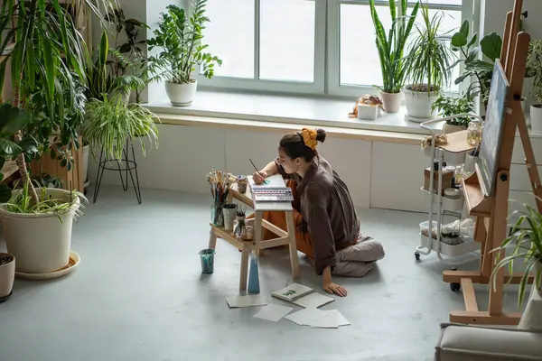 Stock image Creative woman artist sits on floor, painting in cozy art studio with houseplants. Drawing process with inspiration in craft workshop for stress relieve. Imagination, emotions on paper, brush strokes.