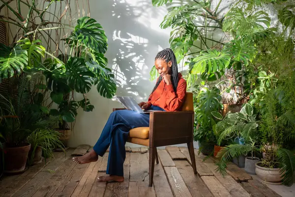 stock image Happy black woman on chair with laptop. African american cheerful girl chatting online using internet sitting among home indoor green garden. Slow life concept, woman resting relaxing in urban jungle.