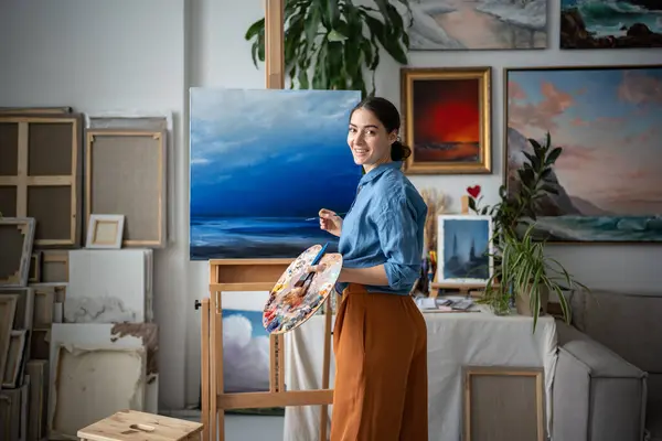 stock image Happy female artist working on oil painting. Cheerful woman painter against of blue picture of sea looking at camera turning around. Creating masterpiece on easel with paints brushes at art studio