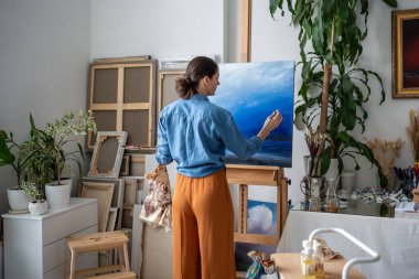 Profesyonel kadın ressam resim üzerinde çalışıyor. Geriye bakış, kadın dikkatlice resim çiziyor fırça, yağlı boya, yaratıcı süreçten huzur hissi alıyor..