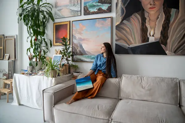 stock image Dreamy female artist sitting on back of sofa, holding painting in hand, looking out window. Woman takes break from creative work, seeking for inspiration, relaxing resting at home art studio.