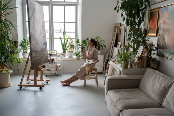 stock image Perfectionist woman artist critically evaluates own painting, sitting on chair, looking for flaws, minuses in work done. Serious strict female painter thinks carefully to refine picture to ideality.