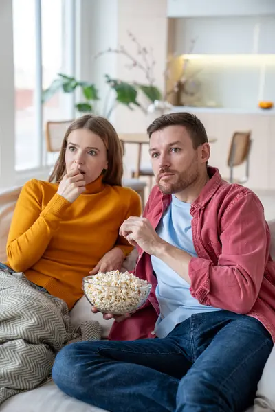 stock image Surprised family couple rest watch TV show, movie, series sitting on couch at home eating popcorn. Amazed relaxed married man and woman enjoy pastime together interested in thriller with exciting plot