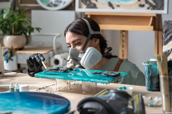 stock image Woman carefully works with wooden stick with epoxy resin, corrects liquid, creating right pattern on canvas. Concentrated female artist in respirator bent close to painting, working out details.