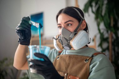 Solunum cihazı ve lateks eldiven takan özenli kadın ressam, sanat stüdyosunda yaratıcı çalışma için mavi epoksi reçine bileşimini test ediyor. Odaklanmış kadın sanatçı deniz manzarası resimleri için sıvı maddeyi inceliyor.