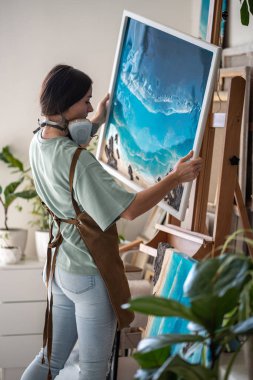 Creative woman artist in apron removed respirator places finished epoxy painting on easel prepare work to be shipped to customer in art studio. Focused girl painter holds painted seascape in workshop clipart