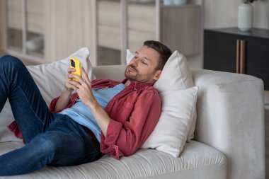 Telefonlu ilgisiz bir adam kanepeye uzanmış internette geziniyor, evde oyalanıyor. Sosyal medya bağımlılığı olan tembel bir erkek akıllı telefondan internete kayıyor, sanal hayatın tadını çıkarıyor..