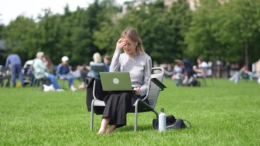 Kulaklıklı tatmin olmuş kadın çıplak ayakla dizlerinin üstünde çalışıyor modern halka açık alandaki çimlerin üzerinde oturuyor. İlgilenen bir kadın ekran bilgisayarına bakıyor. Park çimlerinde video öğreniyor..