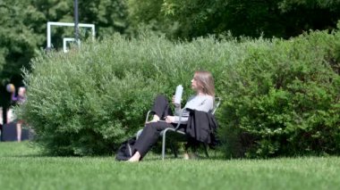 Memnun iş kadını parktaki çimenlerde çıplak ayakla oturur, ofisten sonra sakin vakit geçirir, stresten kurtulmak için temiz havada su içer. Kapalı dizüstü bilgisayarı olan rahat bir kadın, dışarıda çimlerin üzerinde dinleniyor..