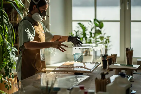 stock image Serious female artist covering finished epoxy resin painting with plastic box, warning canvas against dust, dirt. Woman attentive, careful to created masterpieces. Girl protecting artwork with lid cap