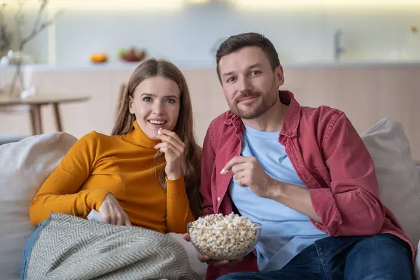 stock image Pleased boyfriend and girlfriend enjoying watching movie while sitting comfortably on couch at home and eating tasty popcorn. Happy interested loving couple engrossed to TV show spend lazy weekend.