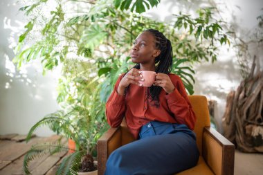 Afro-Amerikalı bir kadının elinde bir fincan kahveyle kenar mahalle ormanlarında bir sandalyede oturup pencereden baktığını hayal etmek. Koltuğunda dinlenen düşünceli siyah kadın ev bitkileri. 