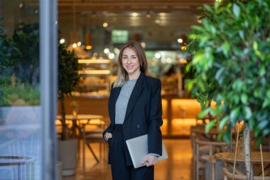 Kendine güvenen ve gülümseyen iş kadını elinde dizüstü bilgisayarla parlak bir kafenin kapısında kameraya bakıyor. İnternet teknolojisinin verimliliğini gösteren bir kadın, küçük işletmeleri ve telekomünikasyonu yönetiyor.