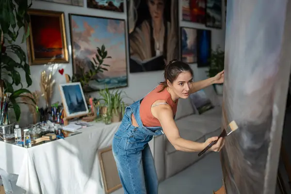 stock image Woman painter crafts landscape on canvas of easel in loft studio. Talented serious female artist working on modern abstract painting. Contemporary art, custom works of professional painter. 