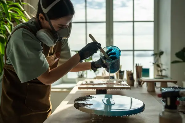 stock image Serious female artist works with epoxy resin, creating picture in technique of liquid art. Professional woman wielding mixing stick, carefully pouring fluid onto canvas. Girl fills artwork with resin