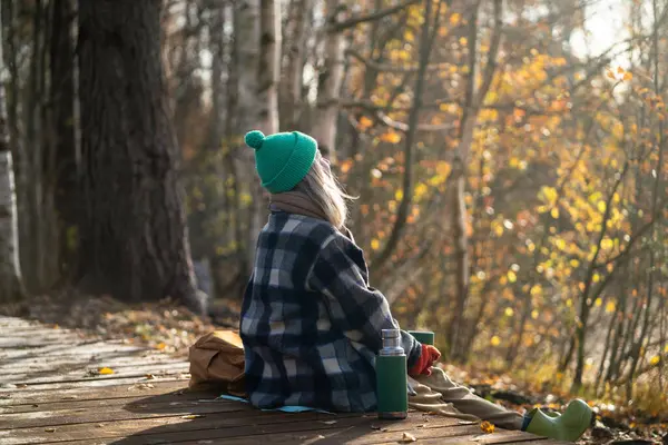 stock image Rearview of serene woman backpacker meditating on eco trail, watching natural landscape in sunny fall woodland. Calm female enjoy autumn air, basking in sun, drinking hot thermos tea in park