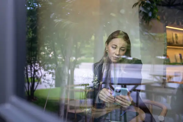 stock image View through glass interested female scrolling news social media feed on smartphone rest in cafe with cup. Businesswoman have coffee break using mobile phone surfing internet web enjoy coffee.