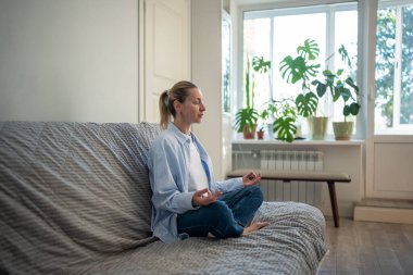 Gözleri kapalı sakin bir kadın Padmasana Lotus pozuyla kanepede derin derin nefes almaya çalışıyor. Yoga ve Mudra egzersizleri yapan meditasyon yapan kadın duygusal dengeyi bulur, rahat duruşu korur.