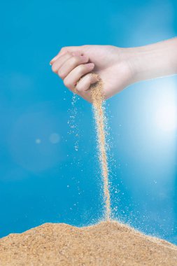 Elle kumları salıyor. Parmaklarının arasından akan ince kum tabakası mavi arka planda. Yaz plajı tatili ve zaman geçirme kavramı. Yüksek hız kepengi izole edildi