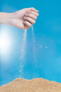 Elle kumları salıyor. Parmaklarının arasından akan ince kum tabakası mavi arka planda. Yaz plajı tatili ve zaman geçirme kavramı. Yüksek hız kepengi izole edildi