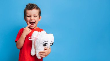 A smiling boy with healthy teeth holds a plush tooth in his hands on a blue isolated background. Oral hygiene. Pediatric dentistry. Prevention of caries. A place for your text