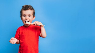 Smiling boy with healthy teeth brushing his teeth with a toothbrush on a blue isolated background. Oral hygiene. A place for your text