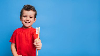 A smiling boy with healthy teeth holds a toothbrush on a blue isolated background. Oral hygiene. A place for your text