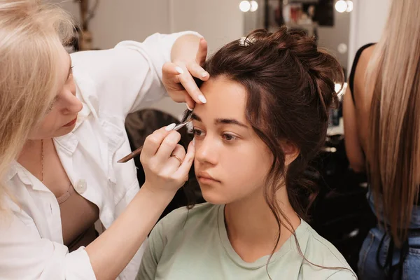 Shooting in a beauty salon. A makeup artist does eyebrow styling for a young dark-haired girl