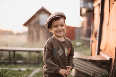 A boy is playing on a farm or ranch field, resting. Portrait of a little boy in a cap and overalls. Childhood. An authentic picture with candid emotions