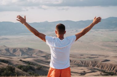 A young man stands with his hands up in the fresh air in a mountainous area
