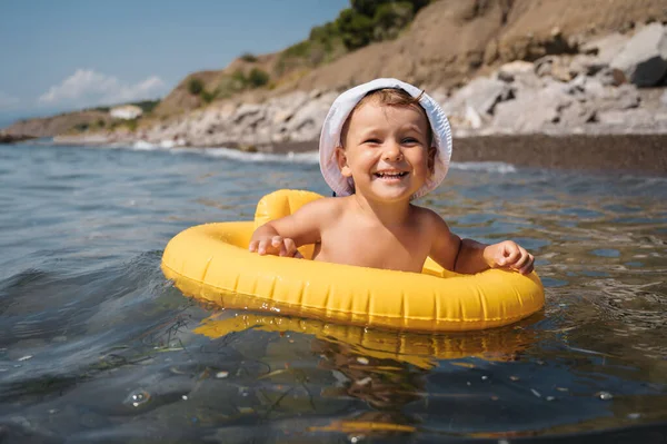 Baby boy swims with an inflatable yellow circle in the sea on a sunny day. The kid learns to swim and enjoys the game