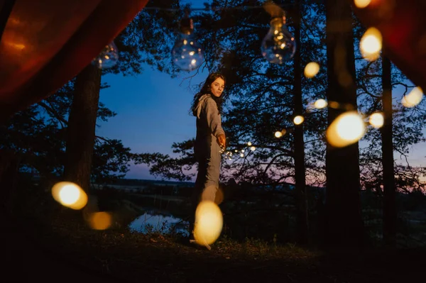 从帐篷里往外看一个年轻的女人站得很高 在树和水的背景下手里拿着灯 户外娱乐活动 魔术就在照片里 — 图库照片